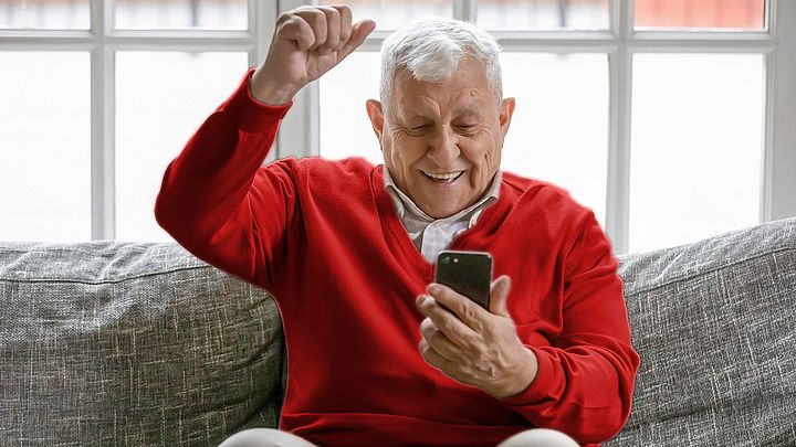 Ein Pensionist hält sein Smartphone in der Hand. Dabei sitzt er auf einem Sofa und ballt vor Freude die Faust.
