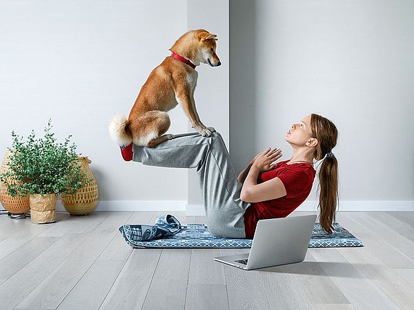 Eine junge Frau macht Sit-ups auf einer Gymnastikmatte in ihrer Wohnung, während ihr Hund auf ihren Beinen sitzt.