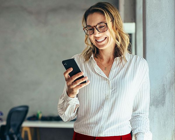 Eine Geschäftsfrau mit Brille steht in einem Büro und blickt dabei lächelnd auf ihr Smartphone