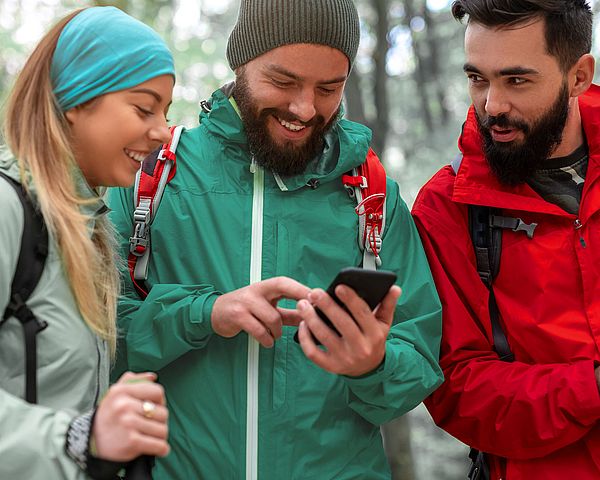 Eine Gruppe glücklicher junger Leute macht beim Wandern einen Halt, um den richtigen Weg zu suchen. Dabei wird das Smartphone als Unterstützung verwendet. 
