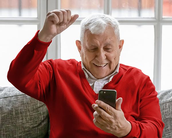 Ein Pensionist hält sein Smartphone in der Hand. Dabei sitzt er auf einem Sofa und ballt vor Freude die Faust.