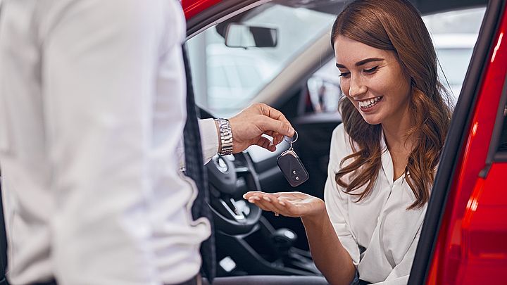 Eine glückliche junge Frau sitzt in ihrem neuen Auto und nimmt den Autoschlüssel vom Verkäufer entgegen.