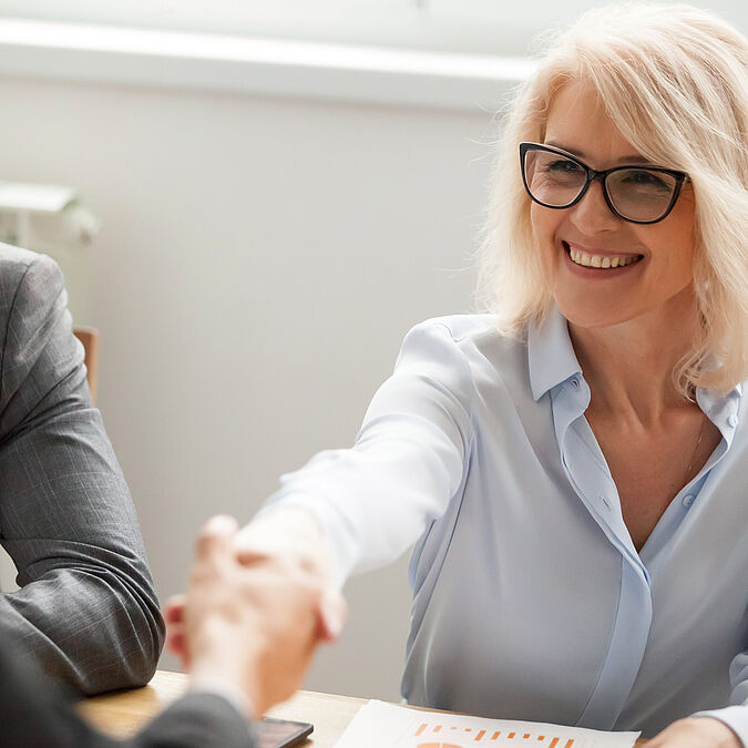 Blonde Frau mit Brille schüttelt jemandem die Hand.