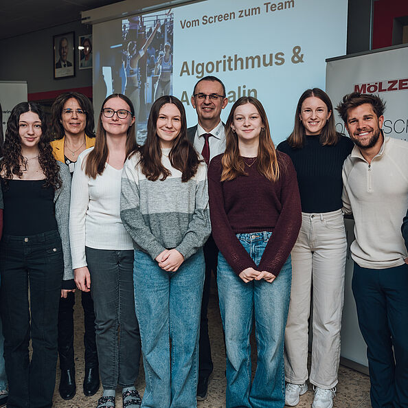 Ein Gruppenfoto mit acht Personen vor einer Projektionsleinwand in einem Raum.