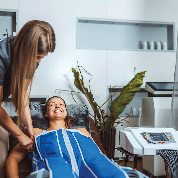 Eine Frau liegt in einem blauen Massageanzug und lächelt, daneben steht eine andere Frau mit langen Haaren, die ihr hilft.