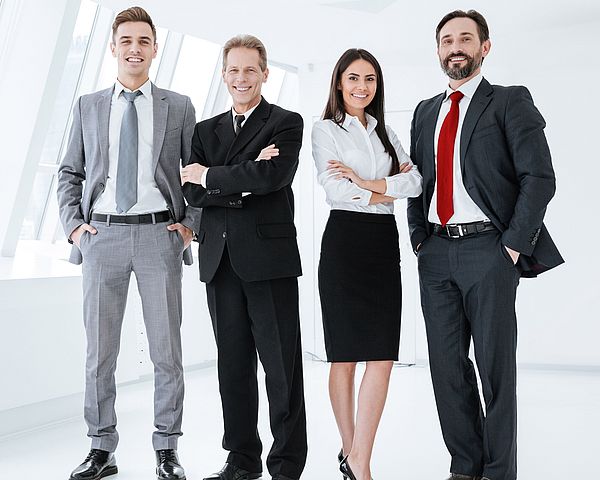 Eine Gruppe von Geschäftsleuten steht in einem Büro zusammen und schauen dabei lächelnd in die Kamera