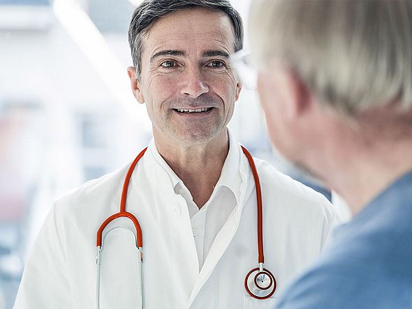 Ein Arzt befindet sich in einer Konversation mit einem älteren Mann und lächelt ihn dabei an. 