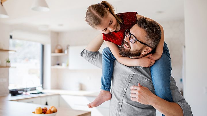 Ein Mann mit Brille trägt zu Hause seine Tochter auf den Schultern und sehen sich dabei direkt an