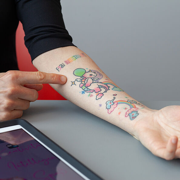 Eine Person zeigt auf ihren tätowierten Unterarm.