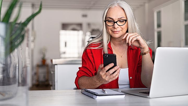 Eine Geschäftsfrau sitzt am Schreibtisch vor ihrem Laptop und hält dabei ihr Smartphone in der Hand, welches sie konzentriert ansieht 