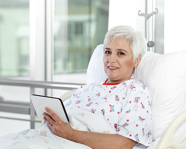 Eine ältere Dame befindet sich im Krankenbett und hält dabei lächelnd ein Tablet in der Hand.