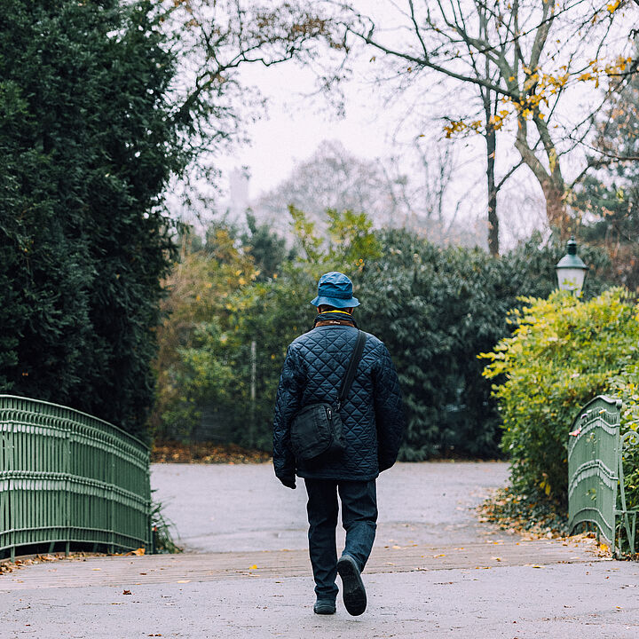 Rückenansicht eines älteren Mannes in dunkler Kleigung, der im Park spazieren geht.
