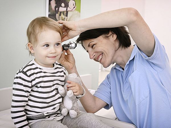 Eine lächelnde Ärztin untersucht ein Kind mit Kuscheltier mit einem Otoskop im Ohr. 