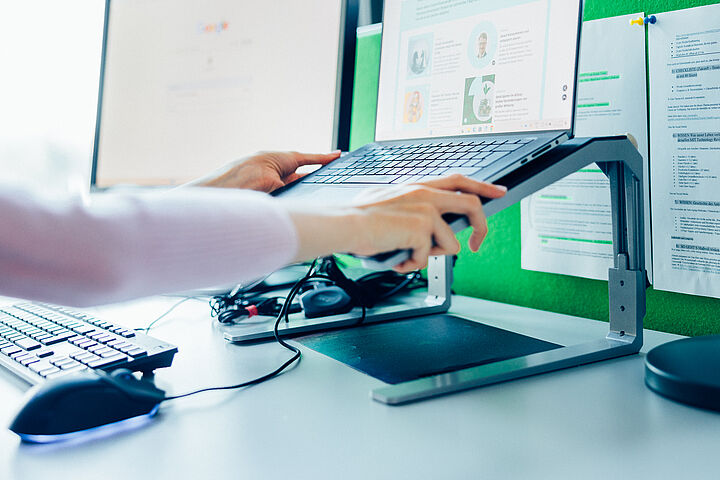 Zwei Hände halten einen Laptop und platzieren ihn auf einem Laptopständer, der vor einer grünen Pinnwand steht, an der zwei Zettel angebracht sind.