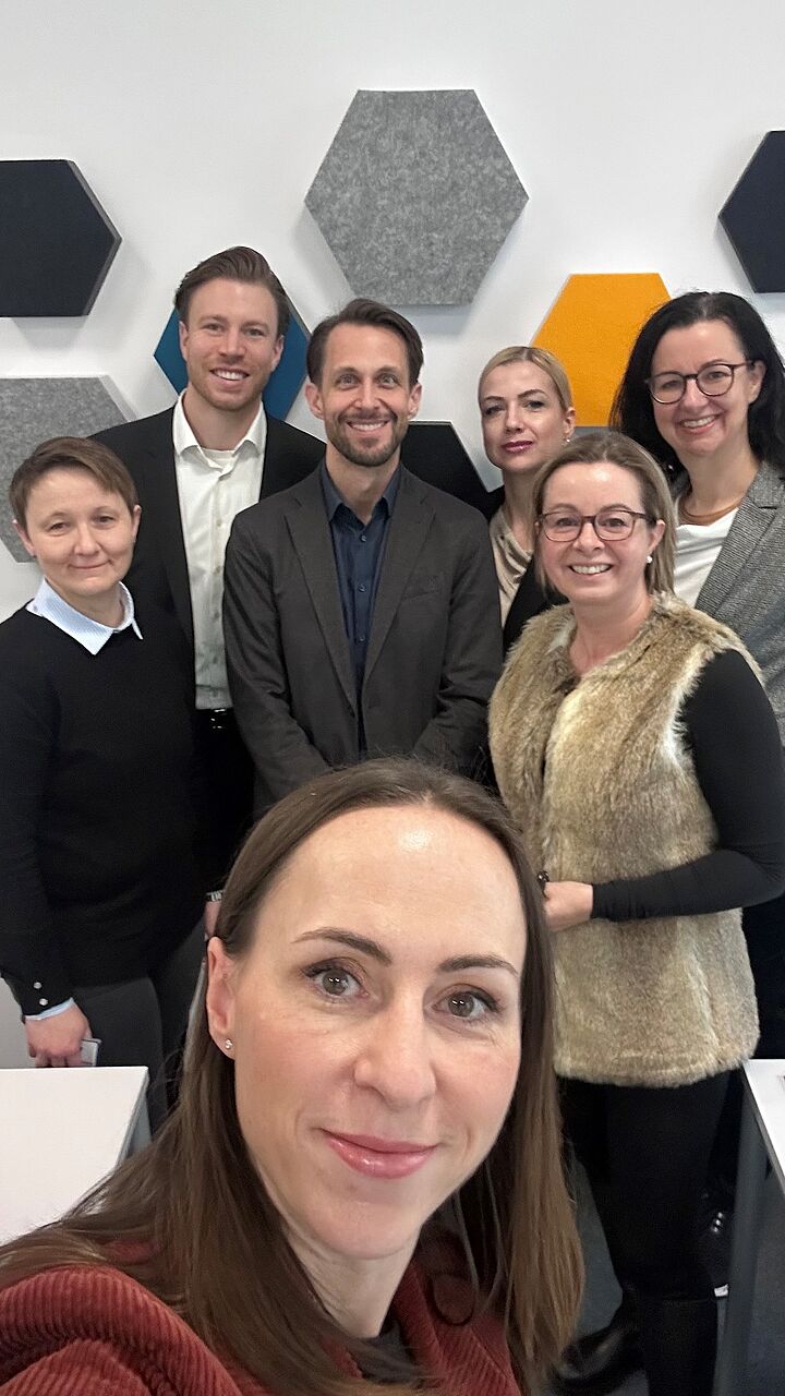 Auf dem Bild sieht man ein Selfie im Bürosetting und 7 Mitarbeiter:innen vor Wand
