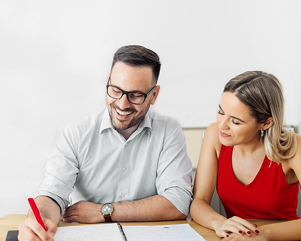 Ein lächelnder Mann und eine Frau sitzen gemeinsam am Schreibtisch, wobei Notizen auf einem Zettel gemacht werden. 