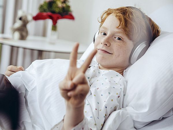 Ein Kind liegt im Krankenbett mit Kopfhörer und zeigt dabei lächelnd ein Peace-Zeichen