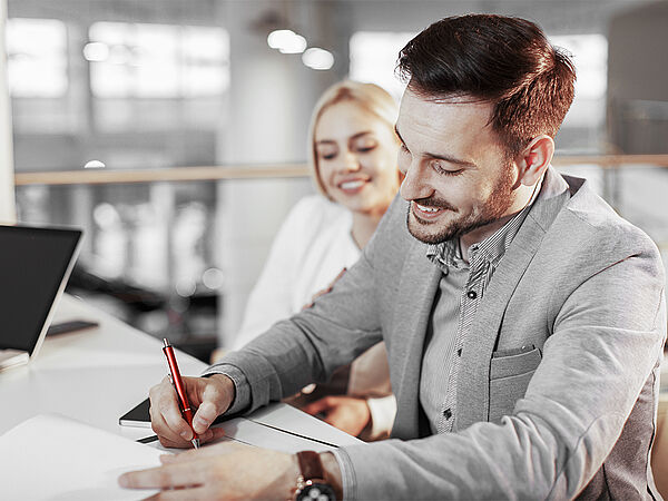 Ein junger Mann unterzeichnet einen Vertrag. Im Hintergrund ist seine Partnerin zu sehen.