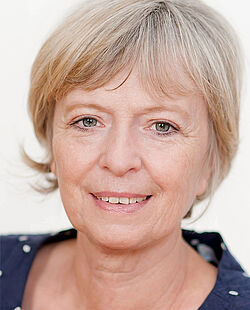 Eine Frau mit kinnlangen blonden Haaren trägt ein dunkelblaues Oberteil mit weißen Punkten und lächelt.