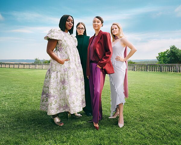 Vier Frauen stehen selbstbewusst nebeneinander auf einer grünen Wiese unter blauem Himmel, elegant gekleidet in moderne, farblich abgestimmte Outfits, mit einer offenen Landschaft im Hintergrund.