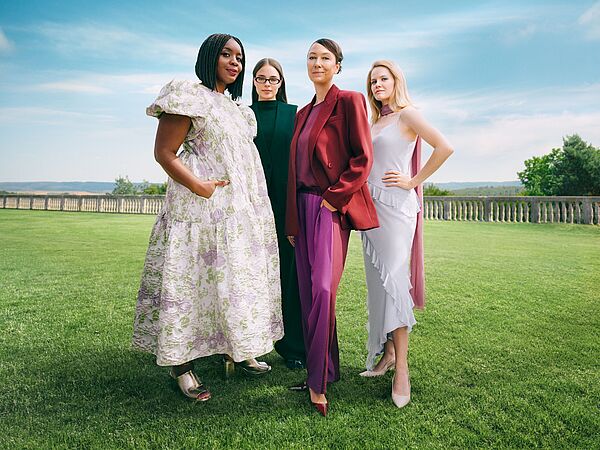 Vier Frauen stehen selbstbewusst nebeneinander auf einer grünen Wiese unter blauem Himmel, elegant gekleidet in moderne, farblich abgestimmte Outfits, mit einer offenen Landschaft im Hintergrund.
