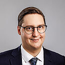 Porträtbild eines jungen Mannes mit Brille, schwarzem Anzug und Krawatte. Er hat kurzes Haar und lächelt.