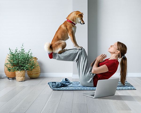 Eine junge Frau macht Sit-ups auf einer Gymnastikmatte in ihrer Wohnung, während ihr Hund auf ihren Beinen sitzt.