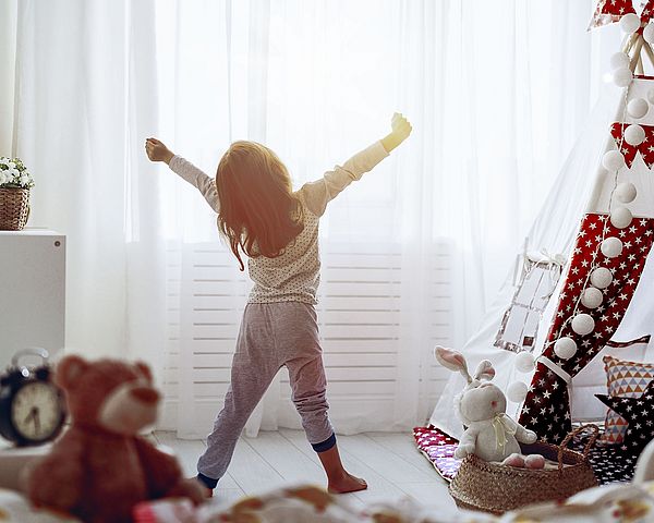 Kind im Pyjama steht vor einem großen Fenster im Kinderzimmer und streckt sich.