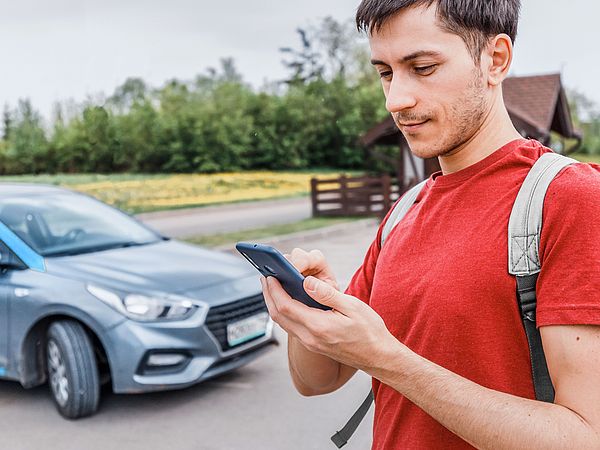 Ein junger Mann steht auf einem Parkplatz neben einem Auto und tippt etwas in sein Smartphone.