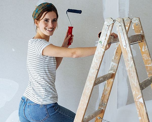 Junge Frau mit Malerwalze lächelt und klettert auf Leiter.