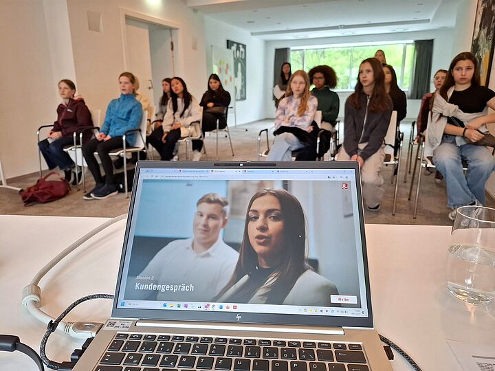 Bildschirm eines Laptops mit geöffneter Präsentation, dahinter sitzen mehrere Mädchen, die dem Vortrag folgen.