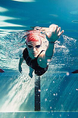 Eine Schwimmerin befindet sich im Becken unter Wasser und schwimmt ihre Bahnen. Dabei hat sie eine Schwimmhaube und eine Taucherbrille auf. 
