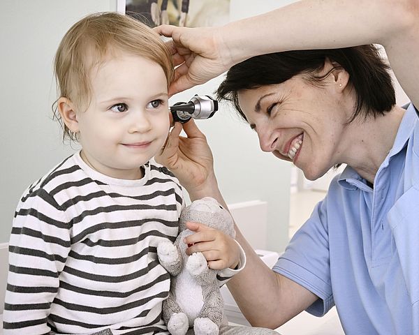 Eine lächelnde Ärztin untersucht ein Kind mit Kuscheltier mit einem Otoskop im Ohr. 
