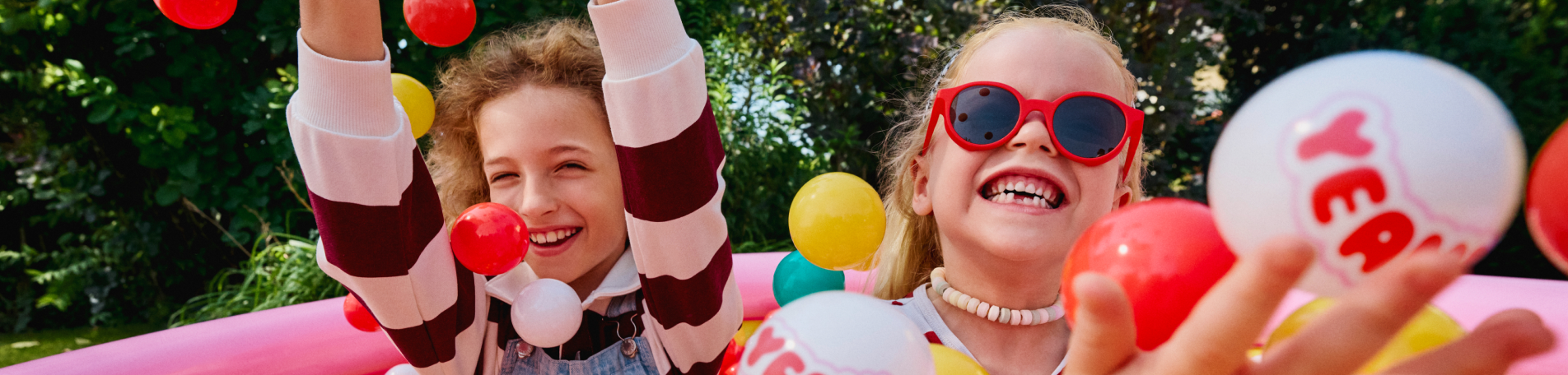 Zwei lachende Kinder spielen in einem bunten Bällebad im Freien und werfen rote, gelbe, weiße und türkise Bälle in die Luft.“