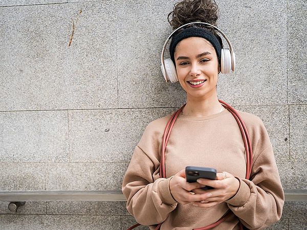 Eine junge Frau mit schwarzen, lockigen Haaren lacht in die Kamera. Sie trägt weiße Over-Ear-Kopfhörer und hält ein Smartphone in beiden Händen.