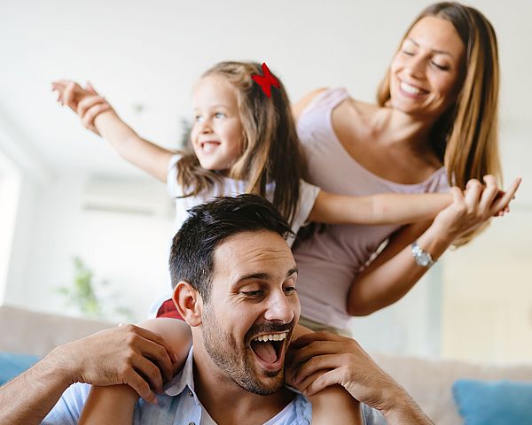 Eine glückliche Familie ist zu Hause im Wohnzimmer. Die Tochter sitzt auf den Schultern ihres Vaters.