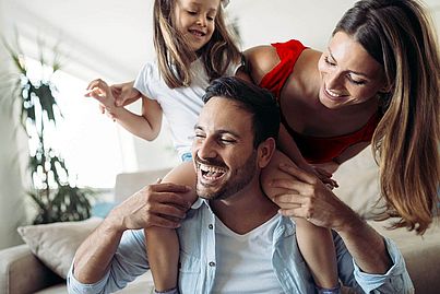 Auf dem Bild sieht man einen Vater mit seiner Frau und seiner Tochter. Beide umarmen ihn.