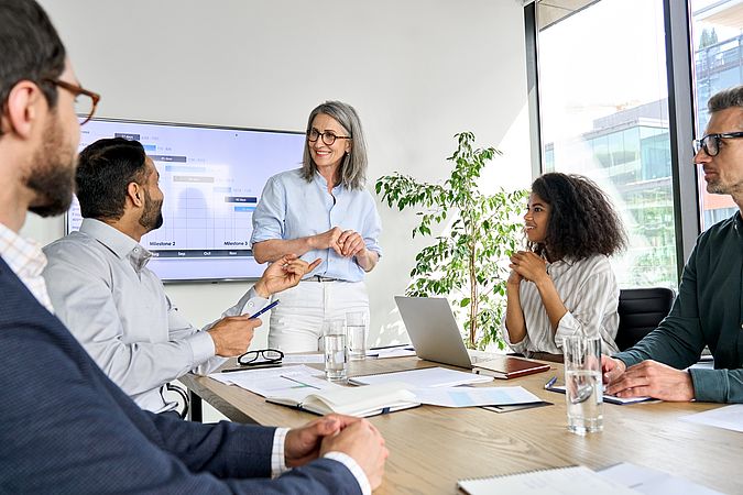 Frau mit Brille steht vor einem Smartboard und hält eine Schulung für vier Mitarbeiterinnen und Mitarbeiter.