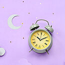 Ein Wecker mit neongelbem Zifferblatt liegt auf fliederfarbenem Untergrund - darüber eine Mondsichel, darunter Wölkchen. Eine Collage.