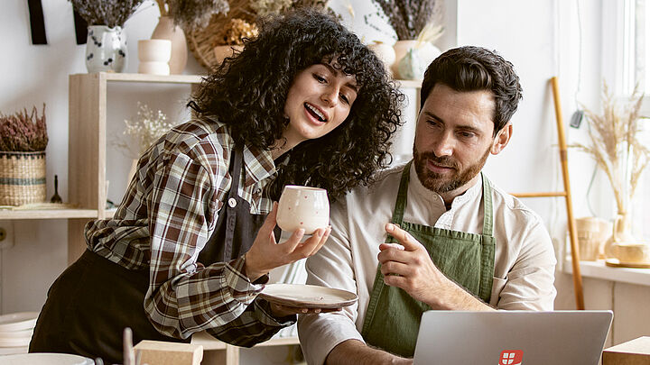 Eine dunkelhaarige, gelockte Frau und ein dunkelhaariger Mann stehen in einer Töpferwerkstatt und betrachten eine selbstgemachte Tasse. Der Mann sitzt vor seinem Laptop.
