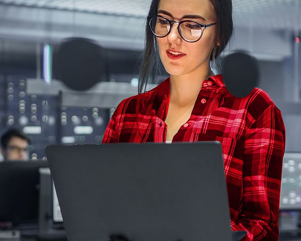 Eine junge Frau befindet sich in einem IT-Büro und arbeitet dabei am Laptop. Im Hintergrund sind Server zu sehen.