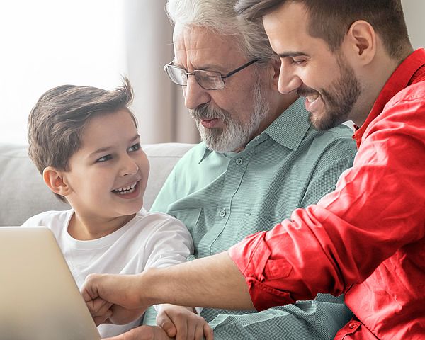 Drei Männer unterschiedlicher Generationen sitzen gemeinsam vor einem Laptop und lächeln sich an.