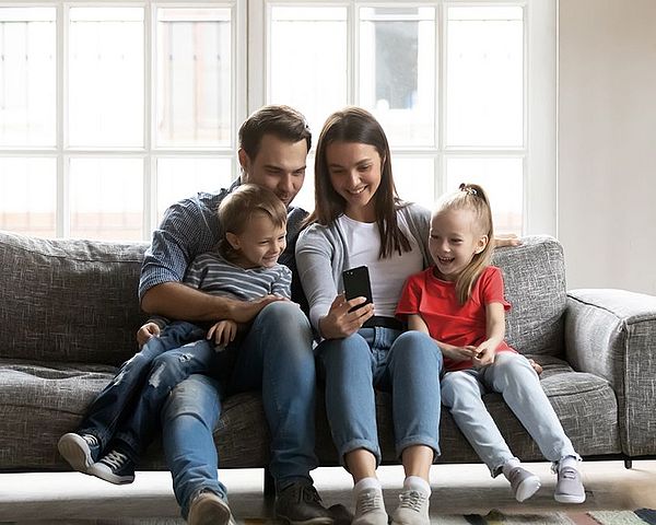 Eine Familie sitzt auf einem Sofa im Wohnzimmer und schaut voller Freude auf ein Smartphone