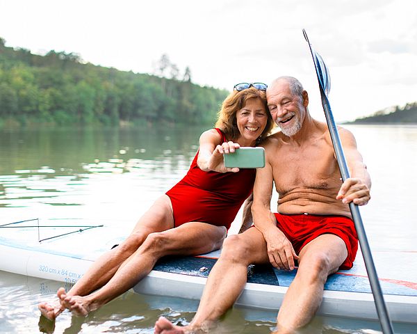 Ein Seniorenpaar sitzt auf einem Paddleboard am See und macht ein Selfie.