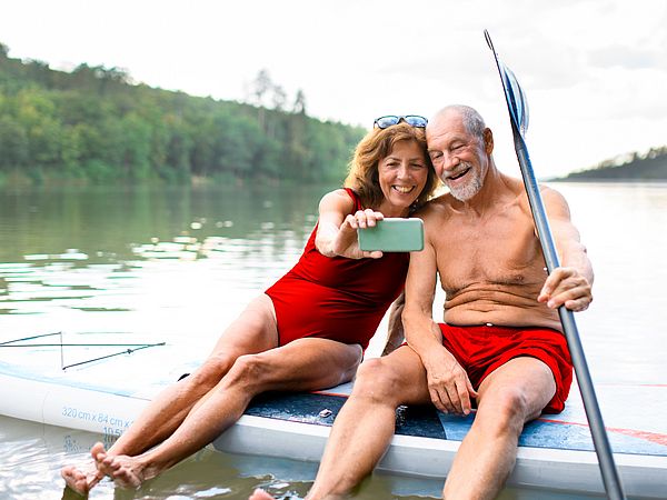 Ein Seniorenpaar sitzt auf einem Paddleboard am See und macht ein Selfie.