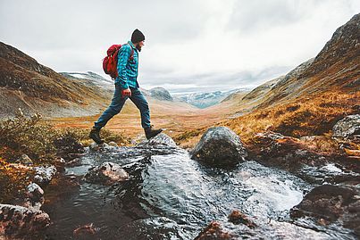 Ein Mann wandert durch eine schöne herbstliche Berglandschaft. Er trägt eine türkise Jacke und einen roten Rucksack.