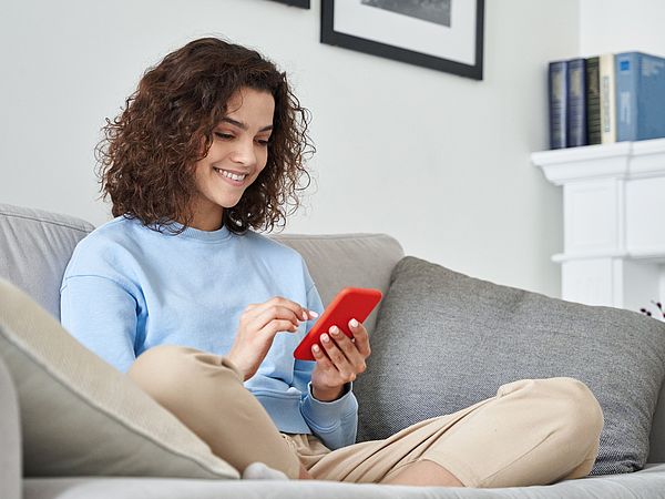 Eine Frau sitzt auf dem Sofa in ihrem Wohnzimmer und tippt dabei auf ihr Smartphone 