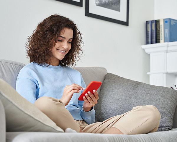 Eine Frau sitzt auf dem Sofa in ihrem Wohnzimmer und tippt dabei auf ihr Smartphone 