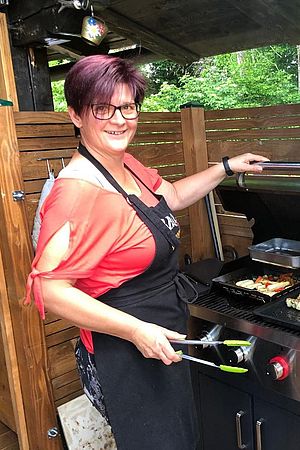 Sandra Schöckenfuchs, eine Außendienstmitarbeiterin der Wiener Städtischen, steht hinter einem Grill in ihrem Garten und bereitet gerade Gemüse zu. Dabei trägt sie eine Schürze und lächelt in die Kamera