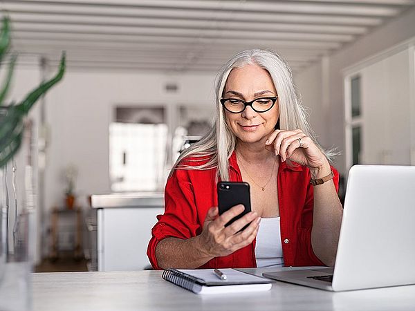 Eine Geschäftsfrau sitzt am Schreibtisch vor ihrem Laptop und hält dabei ihr Smartphone in der Hand, welches sie konzentriert ansieht 